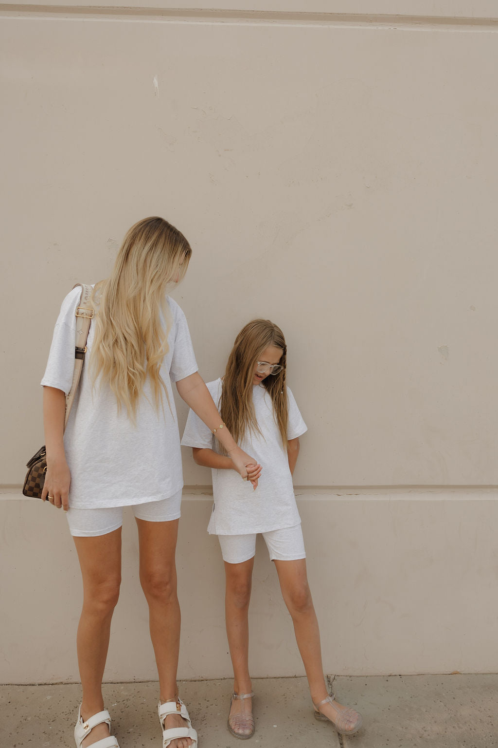 A woman and a young girl, both with long blonde hair, wear matching forever french baby Women's Short Set | Heather outfits as they stand by a beige wall; the woman holds the girl’s arm and looks at her while the girl looks down.