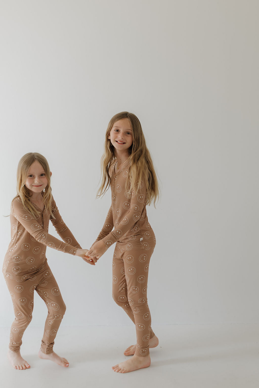 Two young girls with long hair wear matching "Sandstone Smile" bamboo pajamas by forever french baby, holding hands and smiling at the camera against a plain white background.