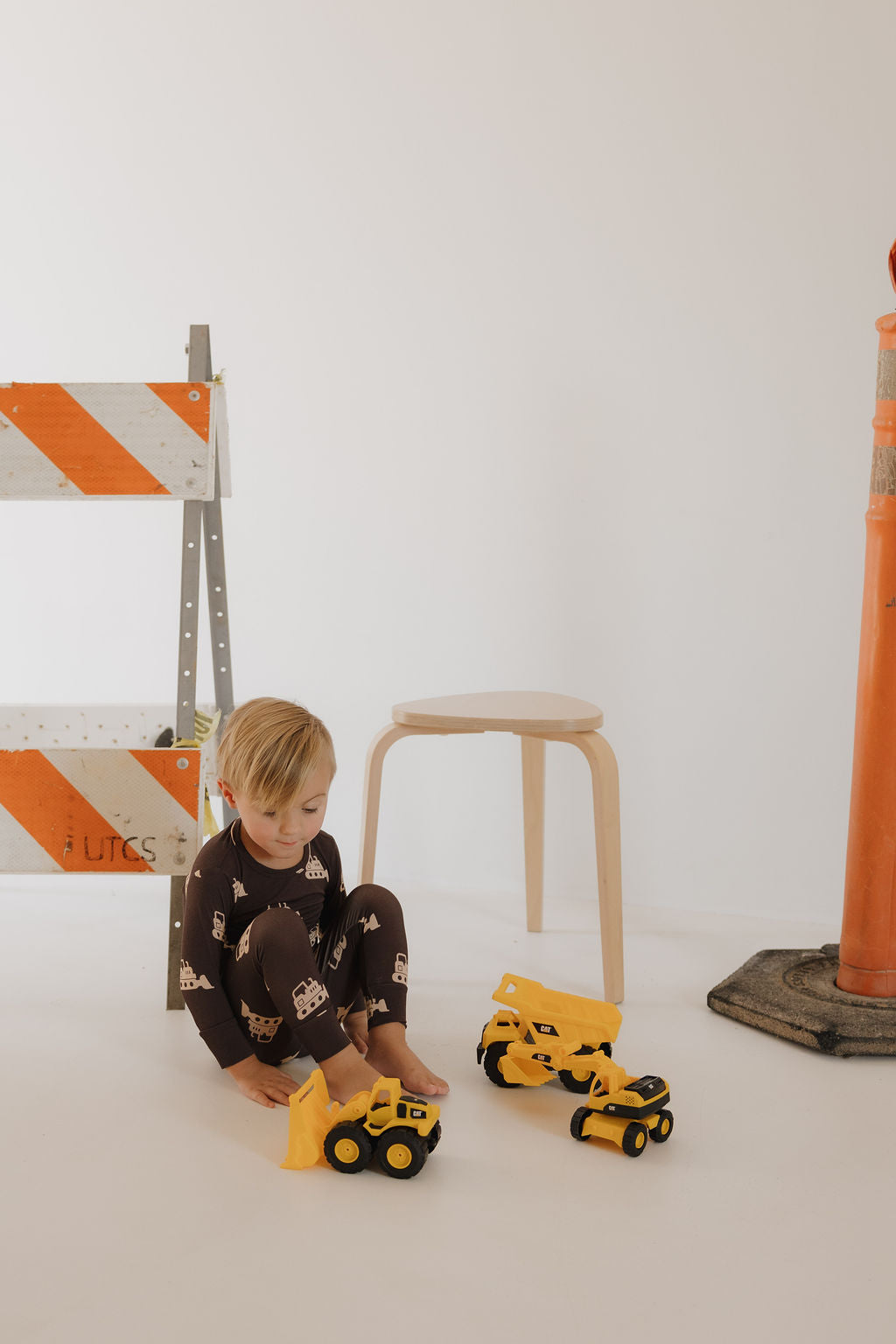 A young child wears the forever french baby Child Sleepy Time Set | Bulldozer pajamas, playing with yellow toy construction vehicles on the floor. Behind him are a wooden stool, an orange safety barrier, and a traffic cone against a white background.