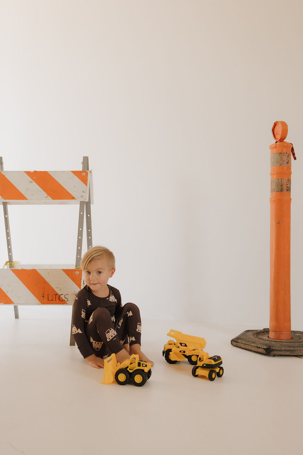 A young child in the forever french baby Child Sleepy Time Set | Bulldozer, breathable brown pajamas with construction vehicle prints, plays with yellow toy trucks by an orange traffic barrier and cone against a plain white background.