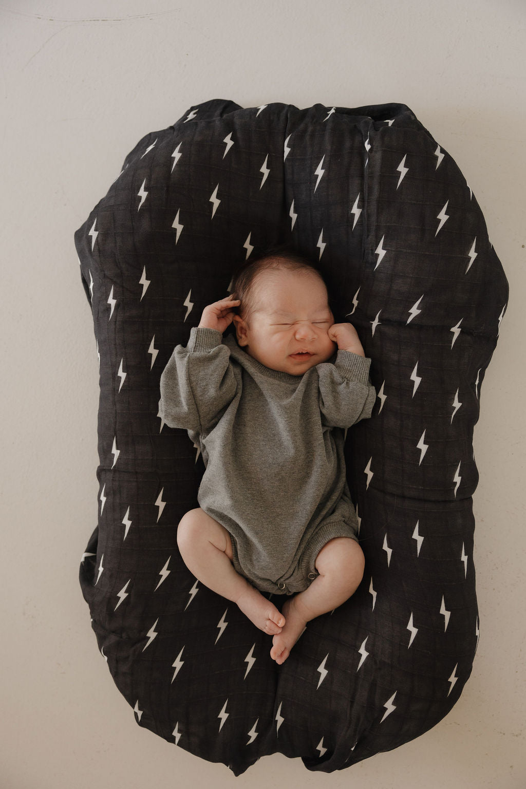 A newborn baby in the forever french baby Sweatshirt Romper | Charcoal lies on a black pillow with white lightning bolts, eyes closed and hands near their face.