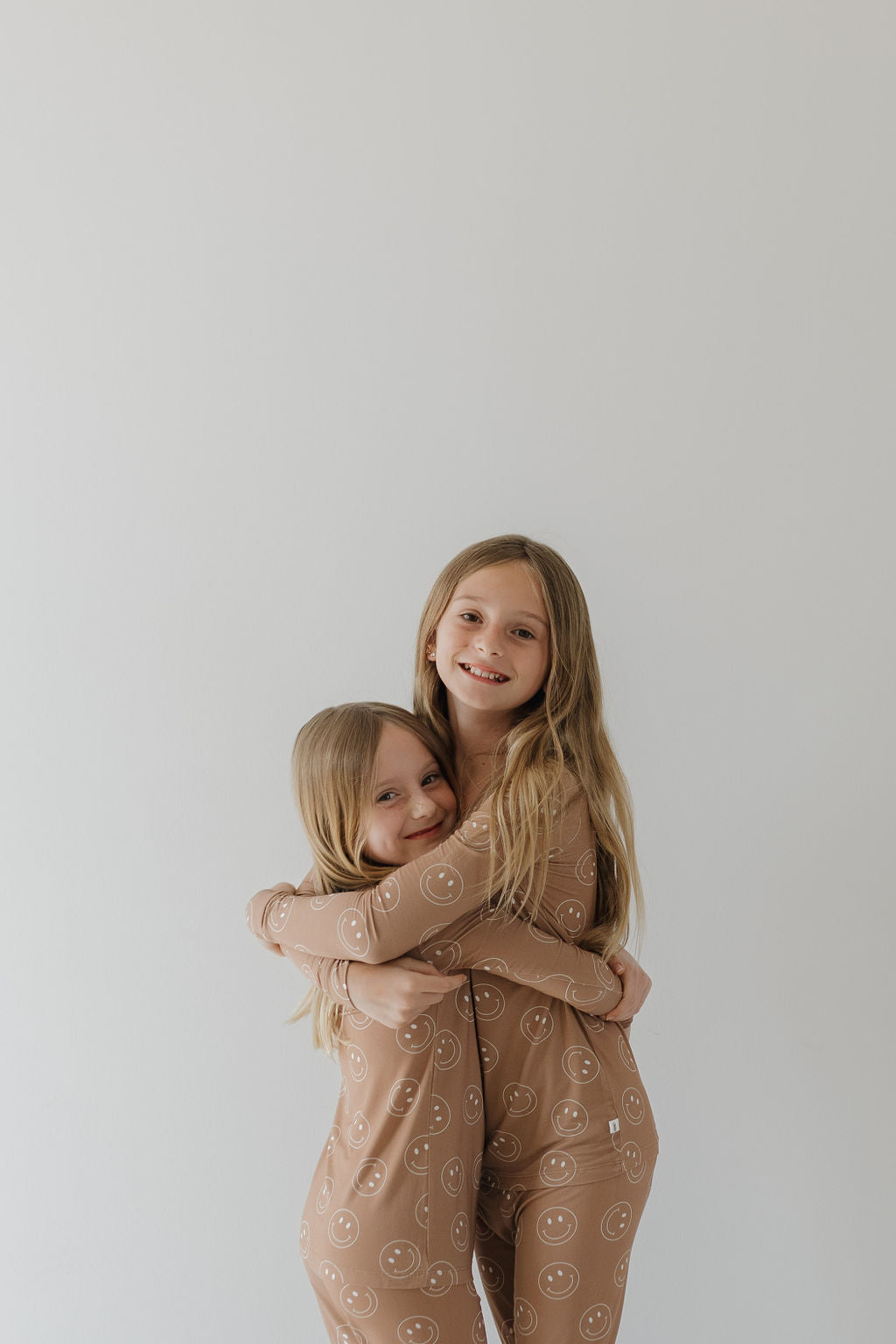 Two young girls with long hair, wearing forever french baby Bamboo Two Piece Pajamas in Sandstone Smile pattern, hug and smile at the camera against a plain light background.