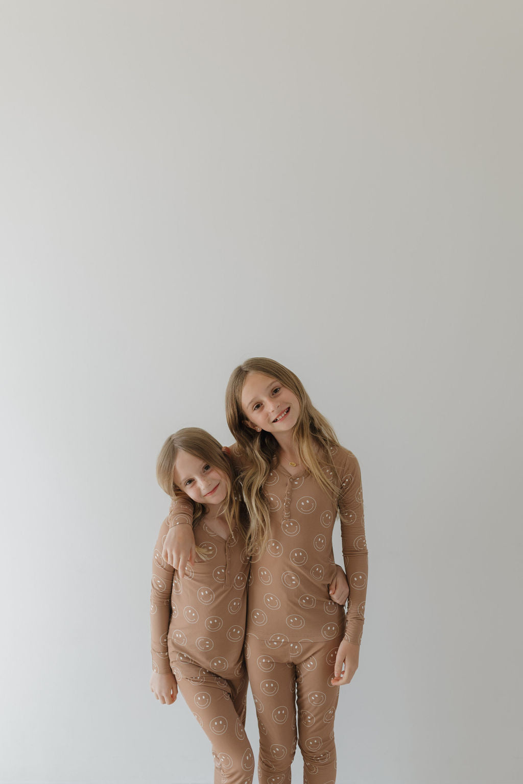 Two smiling children hug in their adorable "Bamboo Two Piece Pajamas | Sandstone Smile" outfits by forever french baby. These hypoallergenic pajamas are crafted from breathable fabric, ensuring comfort against the plain, light-colored background.