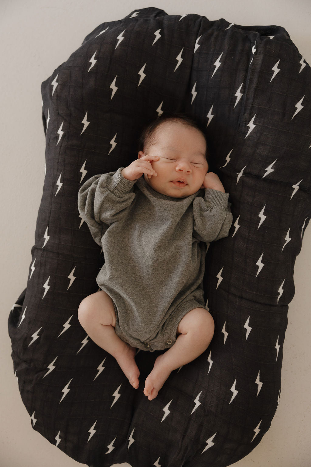 A newborn baby wearing the forever french baby Sweatshirt Romper | Charcoal sleeps on a black cushioned pad with white lightning bolt patterns, arms raised near their face.