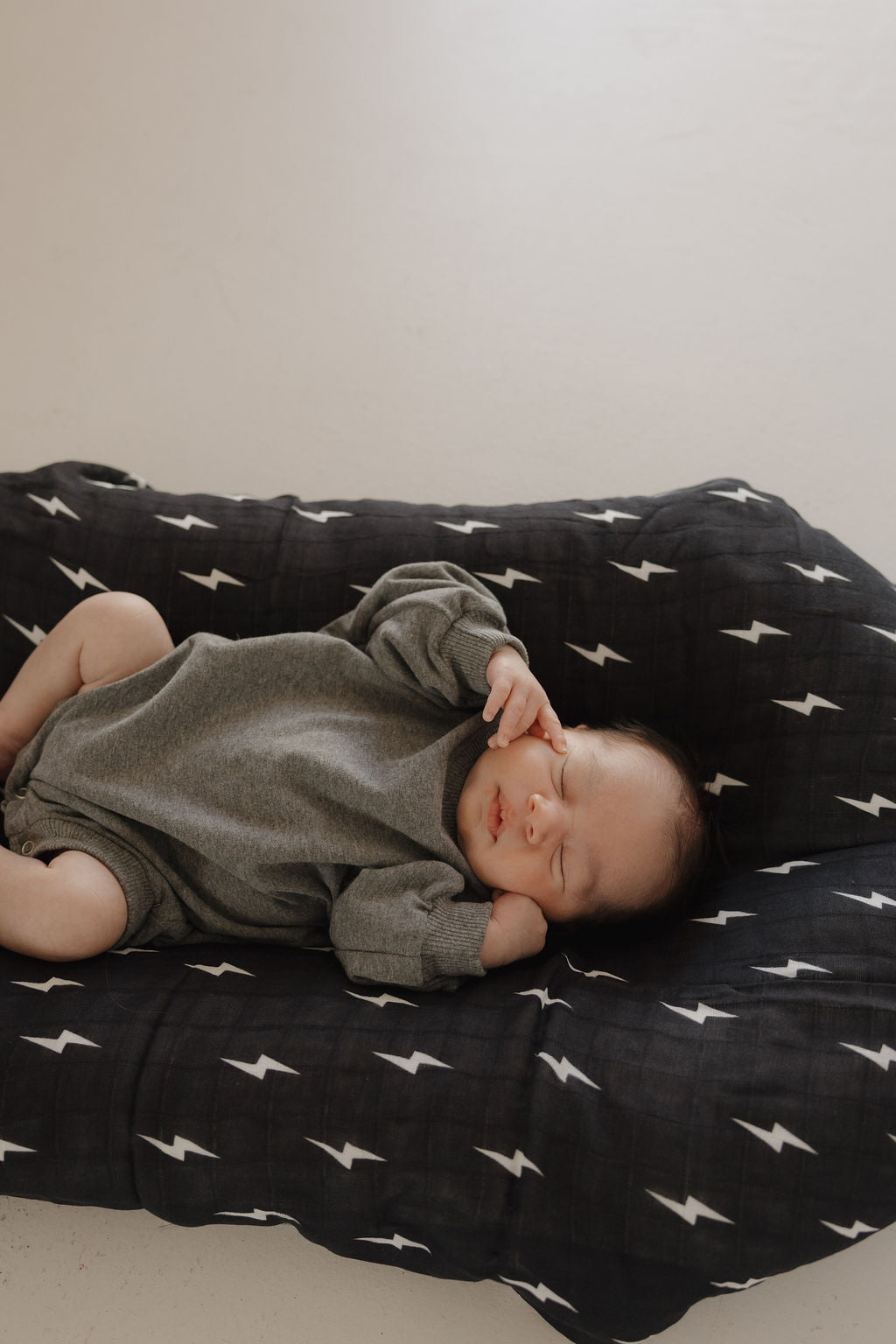 A baby wearing the forever french baby Sweatshirt Romper | Charcoal sleeps soundly on a black cushion with white lightning bolts, hand near their face, against a light neutral background.
