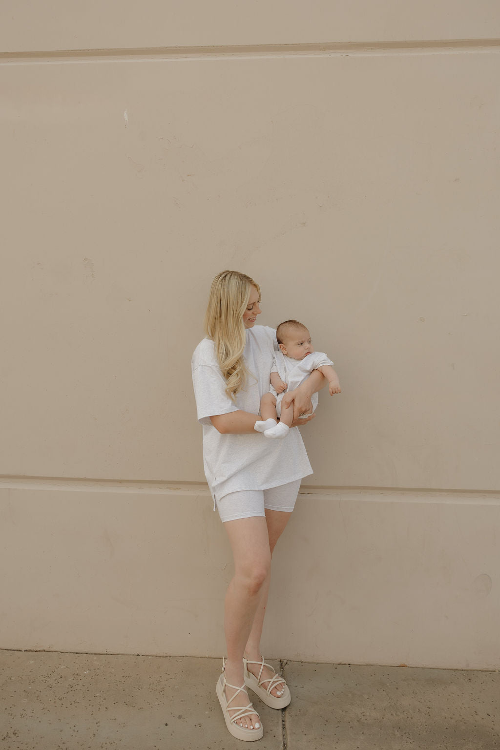 A woman with long blonde hair, dressed in the forever french baby Women's Short Set | Heather, stands against a beige wall holding a baby in white. Both appear content and relaxed.