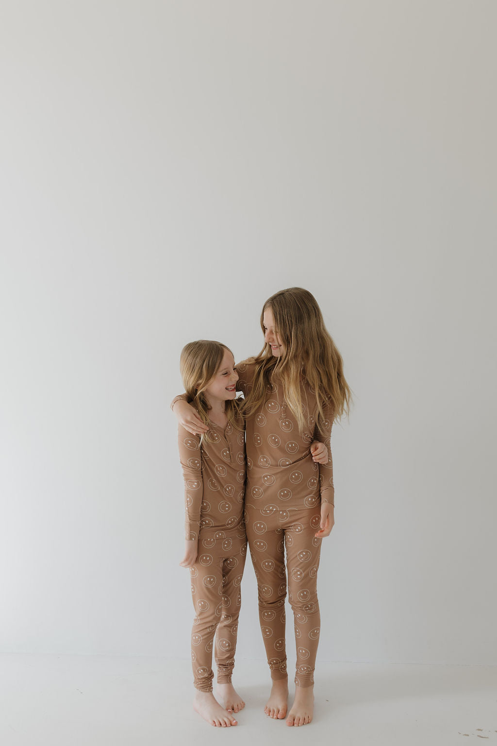 Two children are in matching forever french baby's Bamboo Two Piece Pajamas in Sandstone Smile, barefoot, with one arm around the other's shoulder, smiling at each other against a white background.