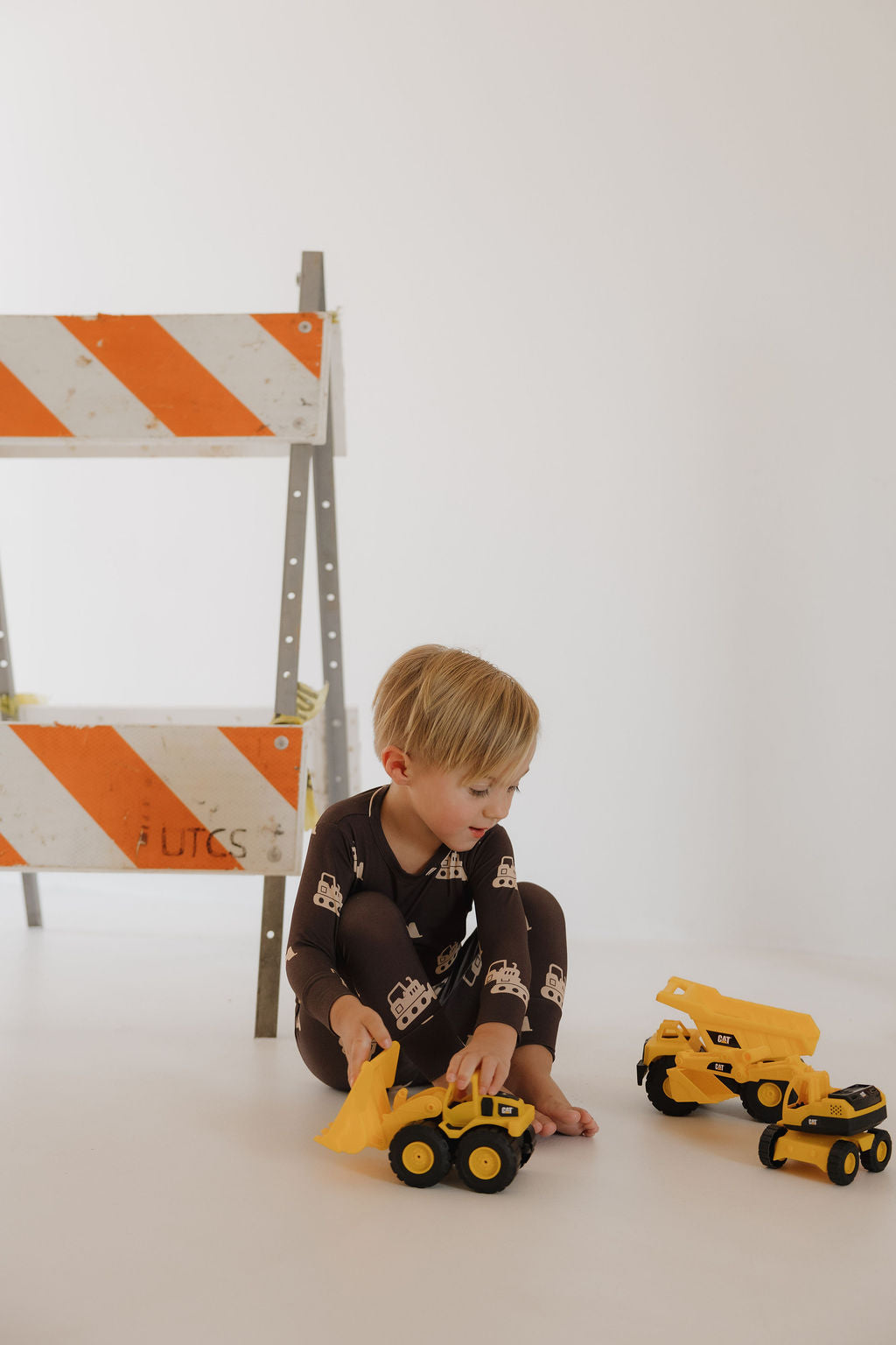 A young child wears the forever french baby Child Sleepy Time Set | Bulldozer—brown, breathable pajamas with construction vehicle prints—while playing with yellow toy trucks beside an orange and white striped barrier.