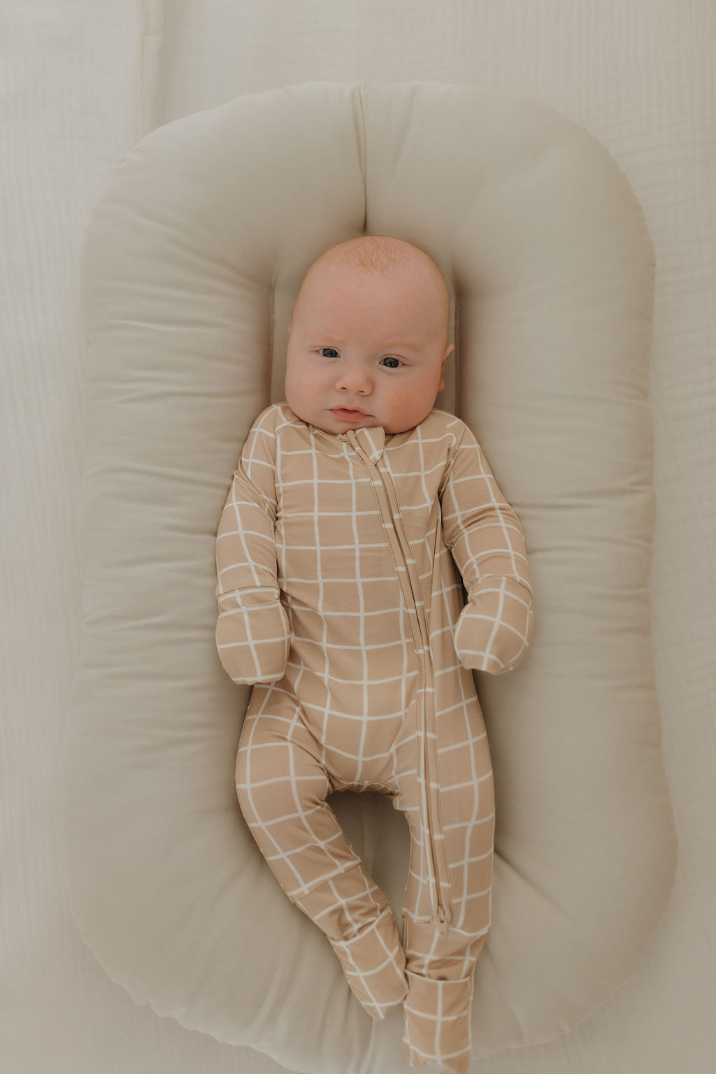 A baby in Bamboo Zip Pajamas | Grid by forever french baby, featuring a tan and white checkered pattern, lies on a soft, light gray cushion. The breathable sleepwear ensures the baby rests in a cozy and peaceful setting, gazing upward with wide eyes.