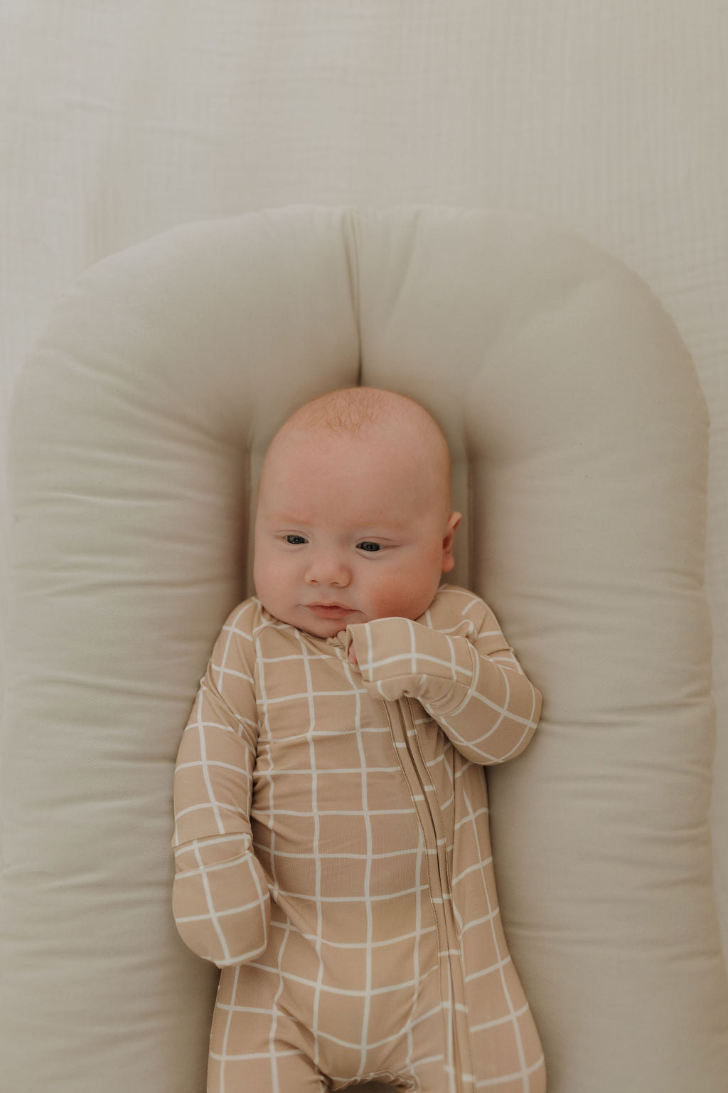 A baby rests on a soft cushion, comfortably wearing breathable Bamboo Zip Pajamas by forever french baby. The beige onesie with a white grid pattern harmonizes with the neutral textured background, while the baby gazes slightly to the side with one hand near their mouth.
