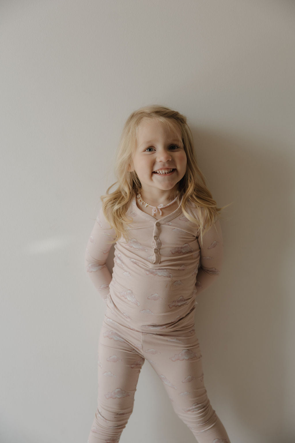A young girl with long blonde hair smiles against a plain wall, wearing Kendy 3’s Bamboo Two Piece Pajamas | Ruby—light pink, hypo-allergenic bamboo pajamas featuring a cloud pattern, with her hands behind her back.