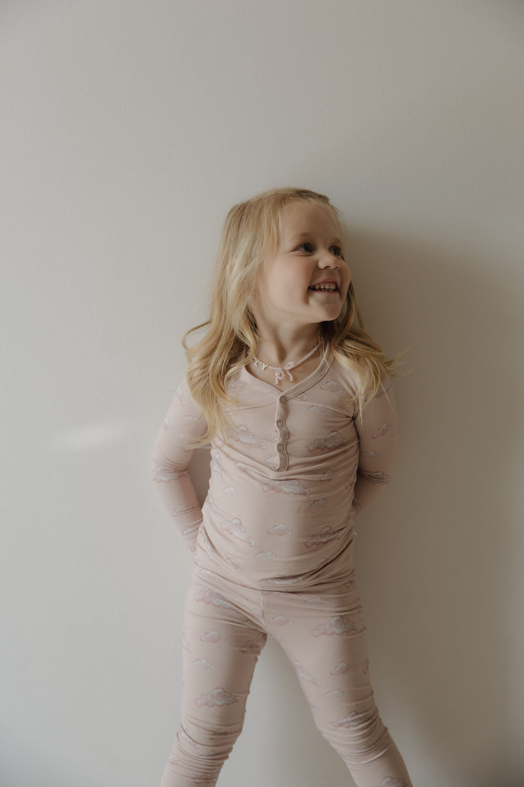 A young child with long blonde hair smiles to the side, hands behind their back, wearing Kendy 3 Bamboo Two Piece Pajamas in Ruby—lightweight and breathable—posing before a plain light wall.