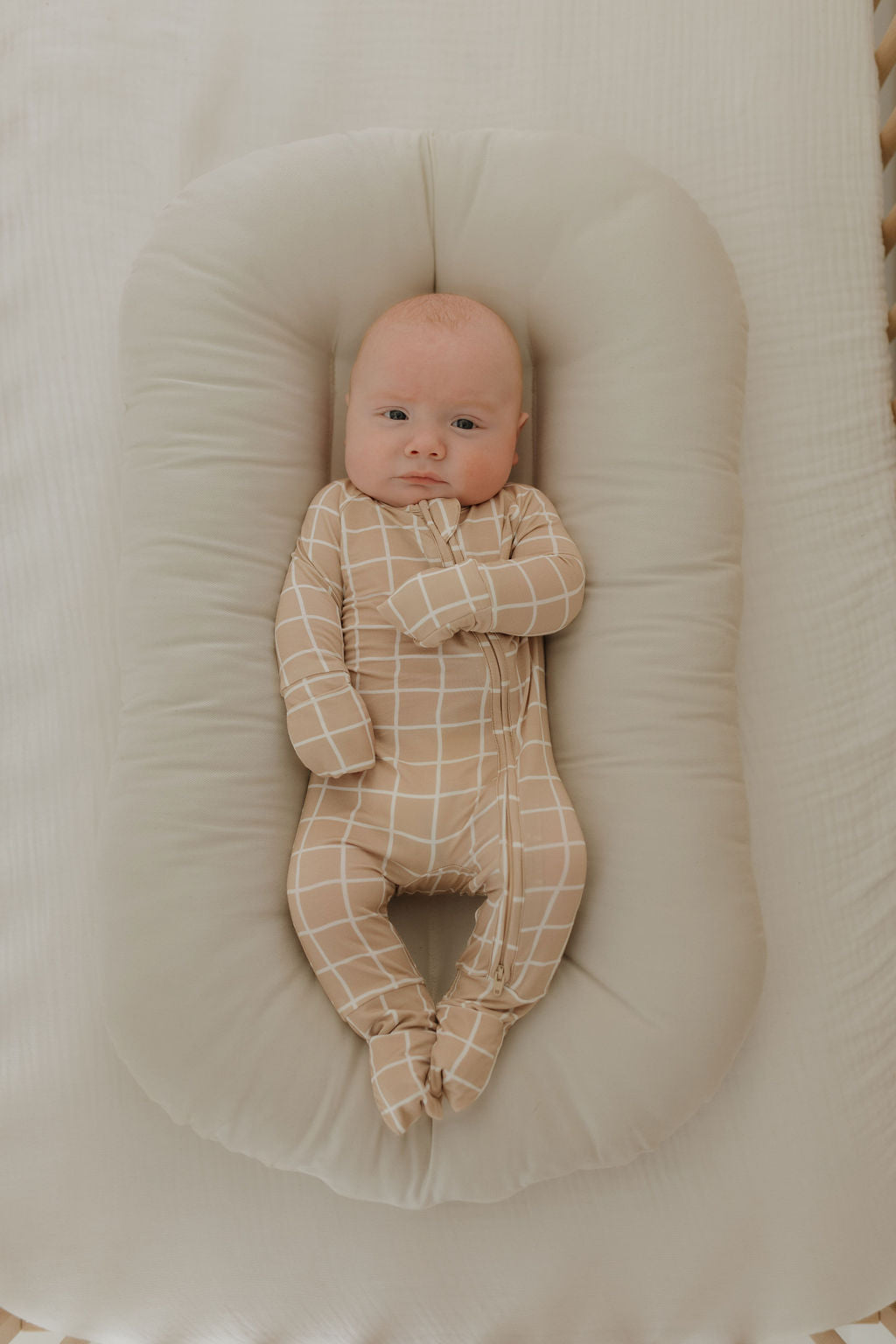 A baby in a beige Grid-patterned Bamboo Zip Pajama by forever french baby lies on a soft, light beige lounger, looking up with a relaxed expression in a cozy, neutral-toned setting.