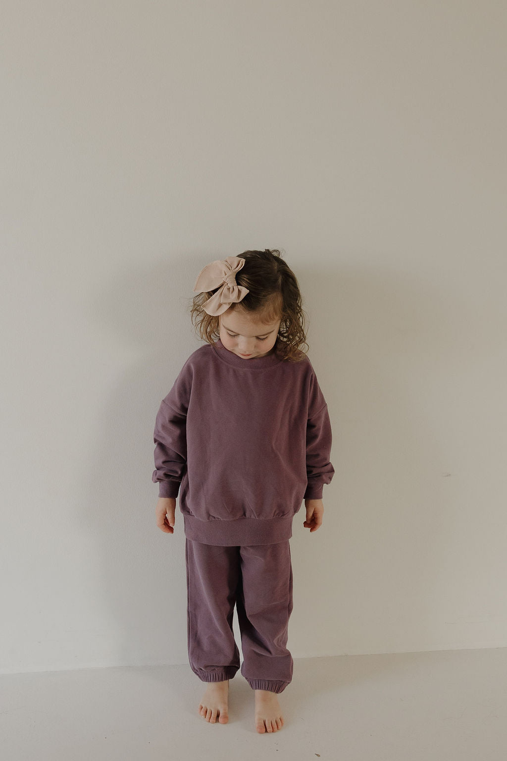 A young child in the forever french baby Child Sweat Set | Twilight stands barefoot by a light wall, looking down. With curly hair and a large beige bow, this outfit is perfect for a coordinated family look.