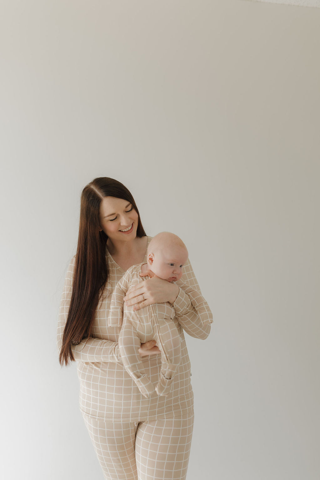 A woman with long brown hair smiles, holding a baby in matching "Women's Bamboo Pajama | Grid" attire from forever french baby. They wear beige checkered, hypo-allergenic clothing against a plain, light-colored wall, enhancing their serene and coordinated style.