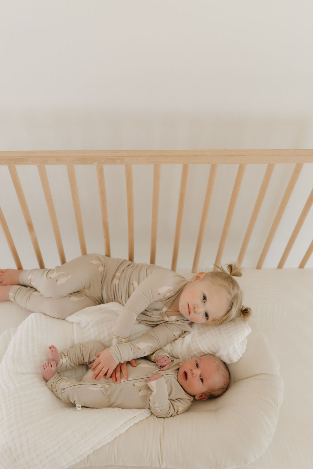 A toddler and baby lie snug on a white bed with wooden rails, both wearing matching Bamboo Zip Pajamas from forever french baby by Silly Goose. The toddler cradles the baby on a soft cushion, portraying a calm and playful scene in their breathable sleepwear.