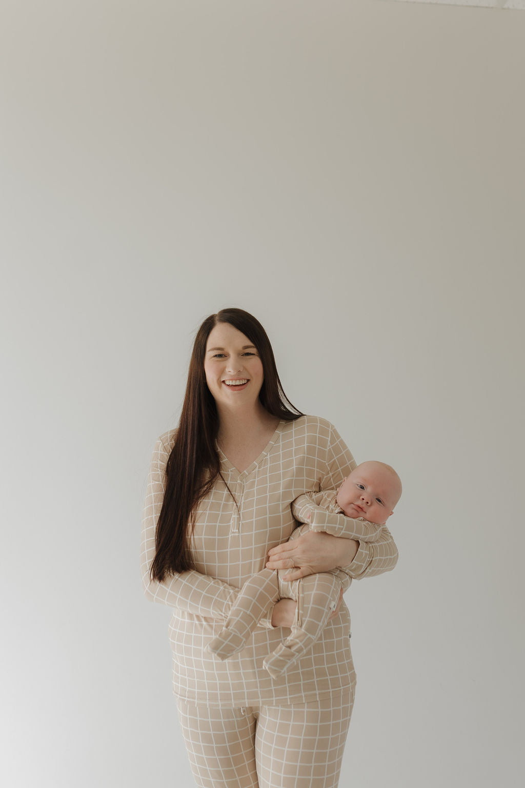 A woman with long dark hair holds a baby, both wearing matching forever french baby hypo-allergenic beige plaid outfits. They stand against a plain light background, smiling at the camera in Women's Bamboo Pajama | Grid attire.