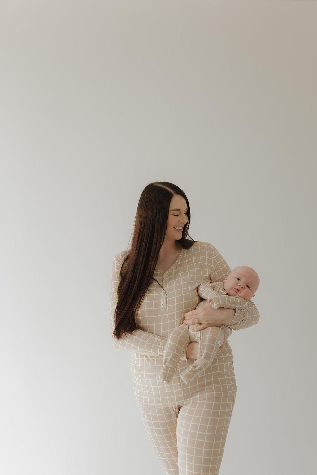 A woman with long dark hair wearing forever french baby Bamboo Zip Pajamas | Grid holds a baby in matching sleepwear. They stand against a neutral light background, smiling warmly at each other.