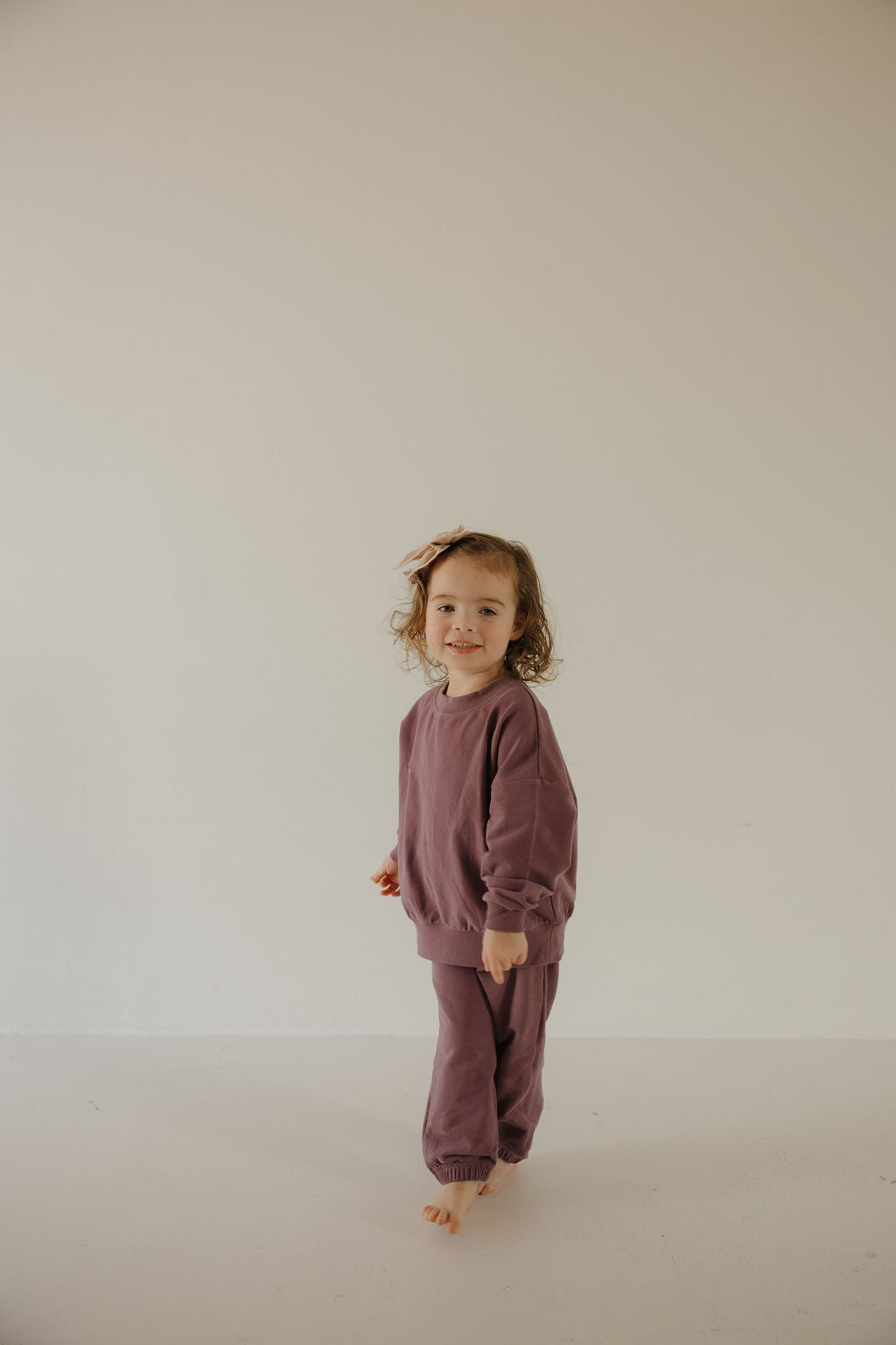 A young child with curly hair wears the forever french baby Child Sweat Set | Twilight, standing barefoot against a plain light background with a gentle smile.