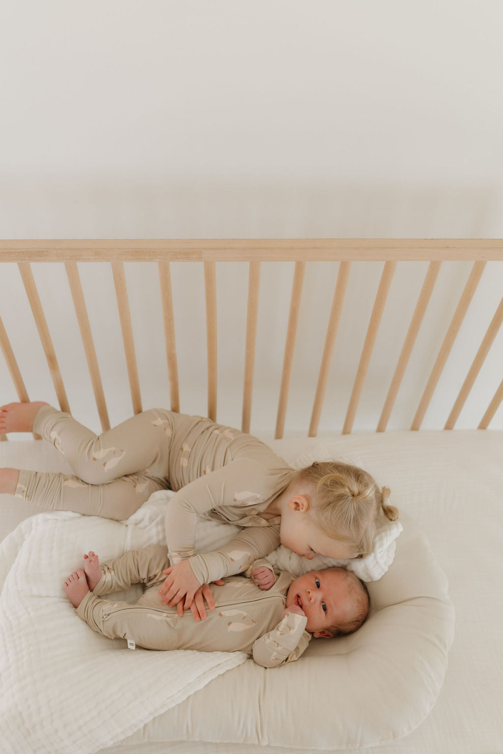 A toddler in matching beige Bamboo Zip Pajamas by Silly Goose kisses a baby lying in a cushioned crib. Both kids are in breathable forever french baby wear, against a plain white background.
