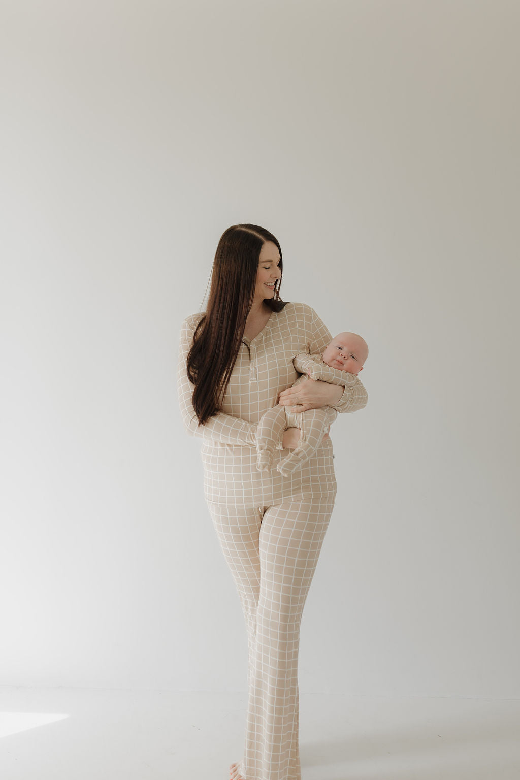 A woman in a matching checkered outfit, the Women's Bamboo Pajama by forever french baby, holds a baby dressed in similar attire. Both are smiling warmly against a plain, light-colored background.