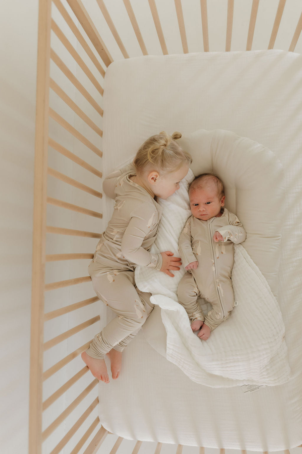 A toddler with blond hair in a bun, wearing light-colored Bamboo Zip Pajamas from Silly Goose by forever french baby, cuddles next to a baby swaddled in matching breathable attire. They lie together in a crib featuring light wooden slats and a white mattress.