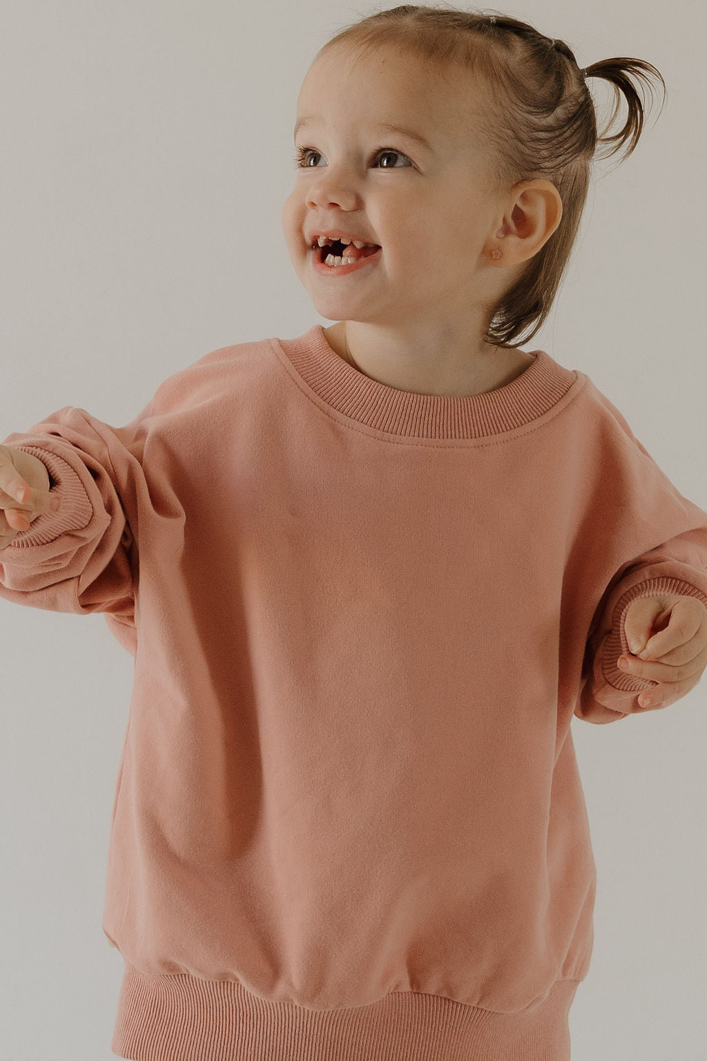 A young child with light brown hair in a ponytail smiles, ready for adventure in the forever french baby Child Sweat Set | Rose, wearing a loose blush pink top against a neutral background.