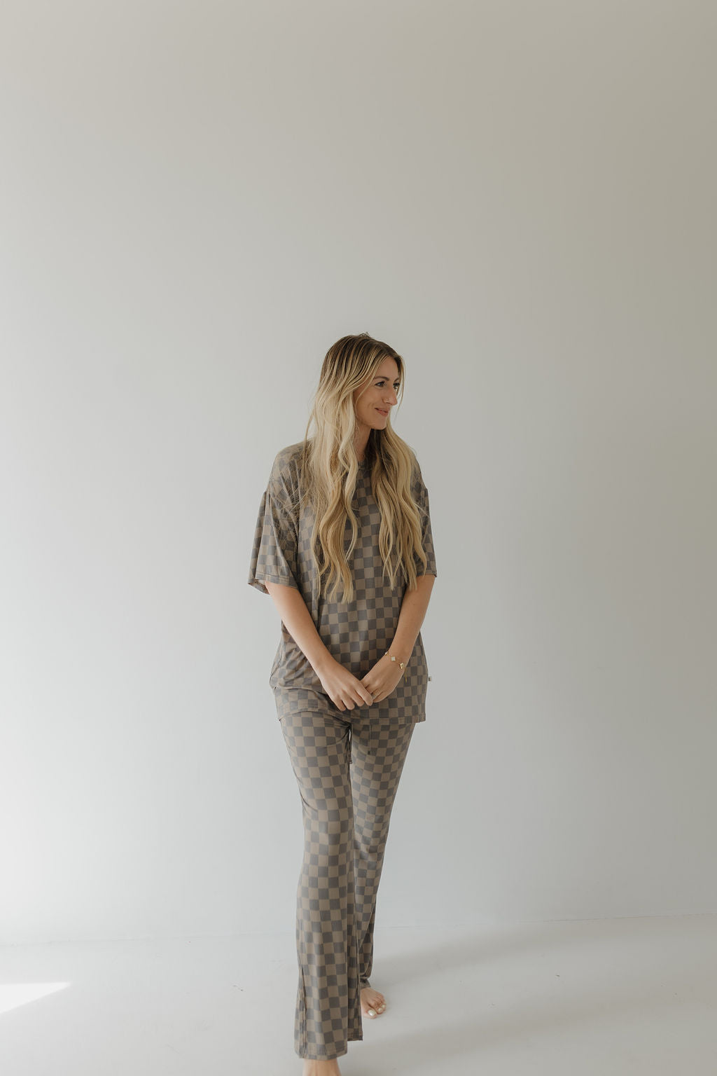 A person with long blonde hair stands against a plain, light background, wearing the Short Sleeve Women's Bamboo Pajama in Faded Checkerboard by forever french baby. The breathable outfit features short sleeves and long pants. They gaze left with hands clasped.