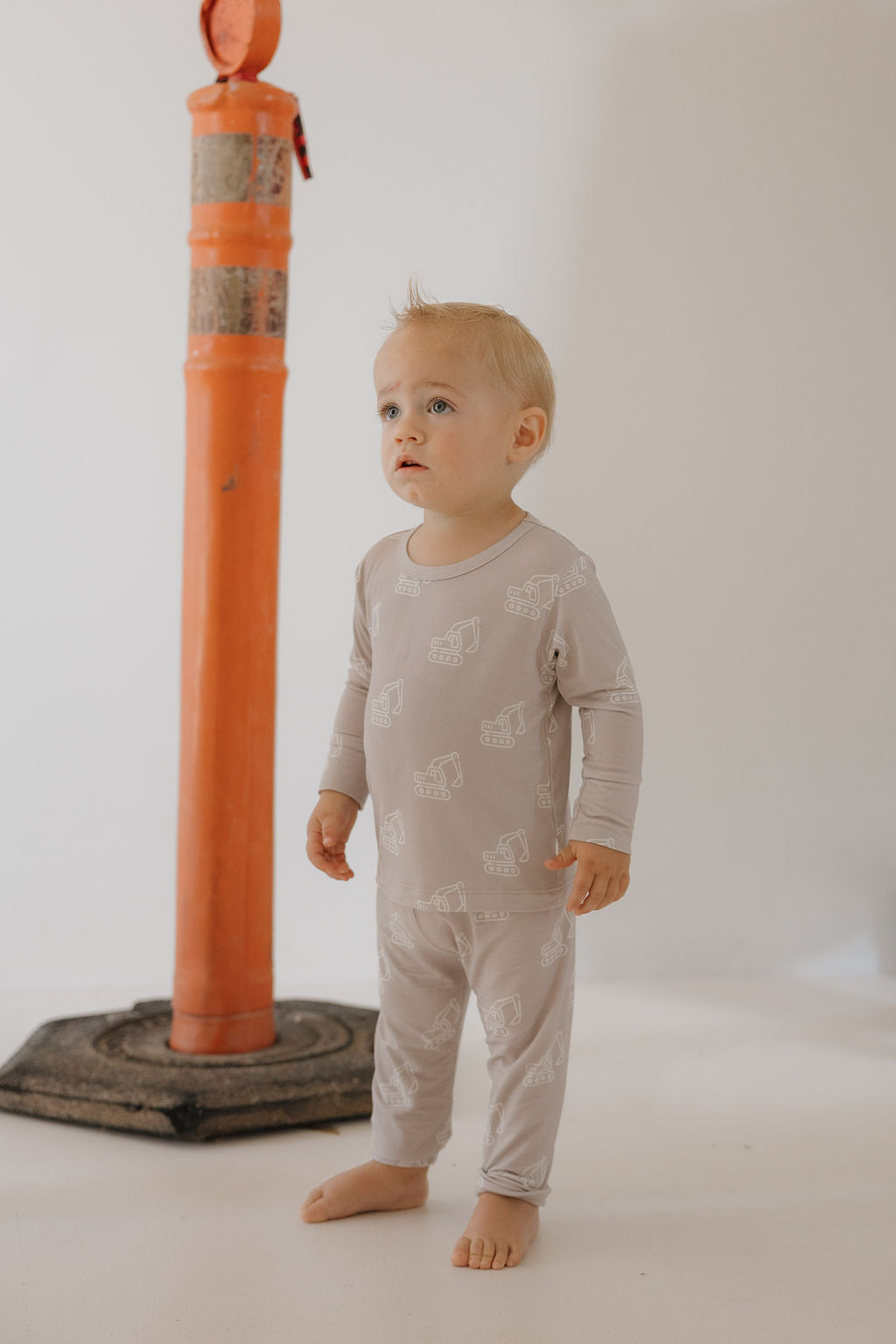 A toddler wears the forever french baby Child Sleepy Time Set | Excavator—light gray bamboo loungewear with white animal prints—standing barefoot on a light floor, with a red and silver pole behind and a plain, light-colored background.