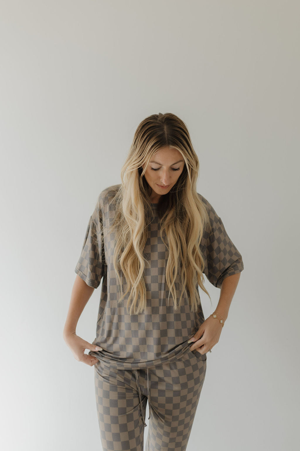 A person with long, wavy blonde hair stands against a plain background, wearing forever french baby's Short Sleeve Women's Bamboo Pajama in Faded Checkerboard. They slightly tilt their head down while casually holding the waistband of the breathable outfit.