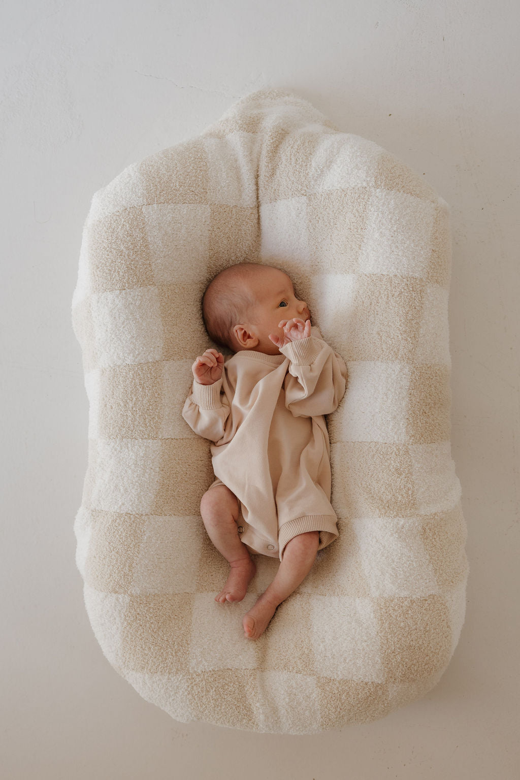 A newborn wearing the forever french baby Sweatshirt Romper in Cashew lies on a soft, beige and white checkered cushion, gazing to the side with one hand near their mouth.