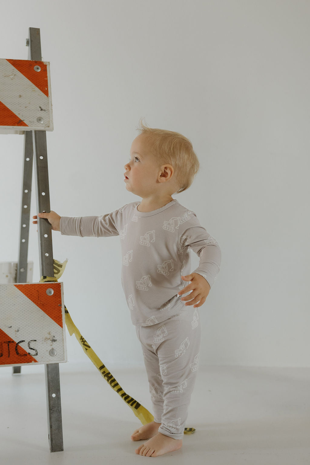 A barefoot toddler wears the forever french baby Child Sleepy Time Set | Excavator, standing indoors beside a metal traffic barricade with red and white stripes and yellow caution tape, looking up and to the side.