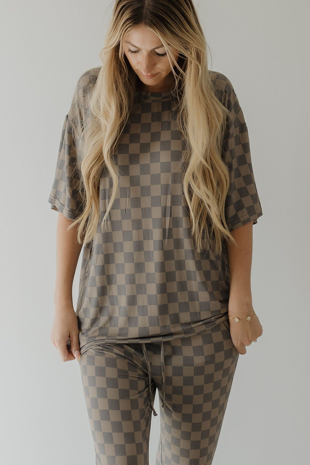 A person with long, wavy blonde hair wears a forever french baby Short Sleeve Women's Bamboo Pajama in Faded Checkerboard. They stand against a plain light background, looking down slightly, with hands resting on their comfy pajamas.