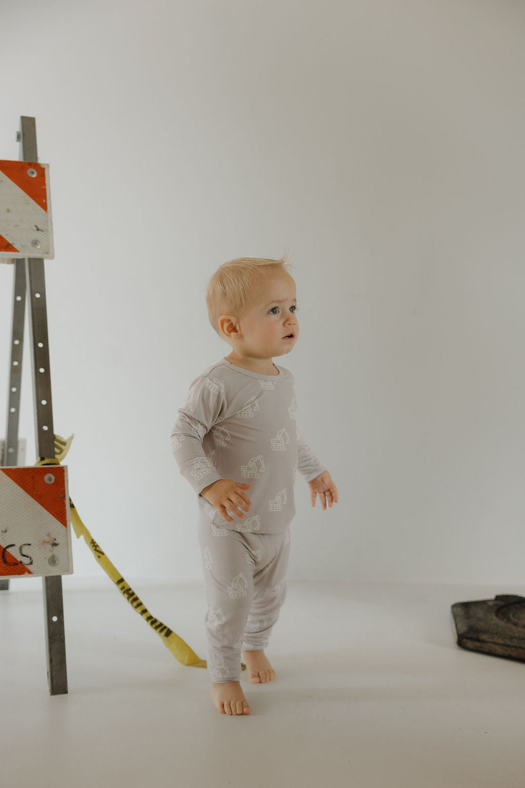 A toddler wearing the forever french baby Child Sleepy Time Set | Excavator—light gray, hypo-allergenic pajamas with white animal prints—stands barefoot indoors by a construction barrier and yellow caution tape against a minimal background.