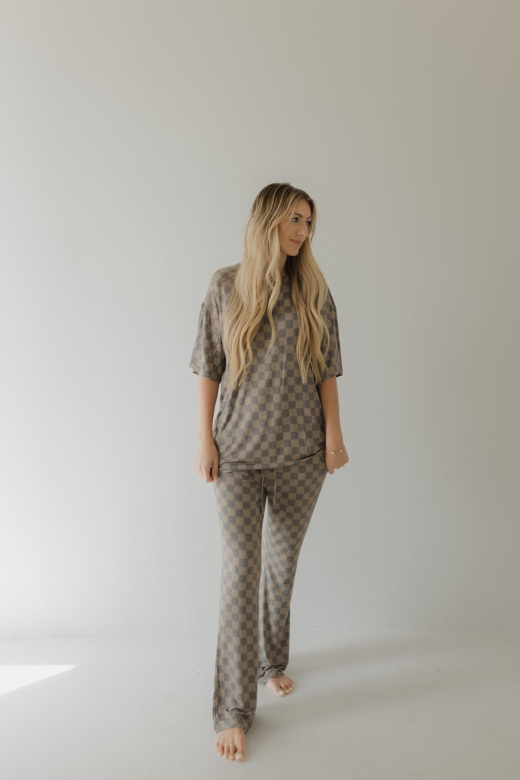 A person with long blonde hair stands barefoot in the Short Sleeve Women's Bamboo Pajama by forever french baby, featuring a faded checkerboard design. The light-colored background highlights the breathable, gentle style of this brown and beige ensemble.