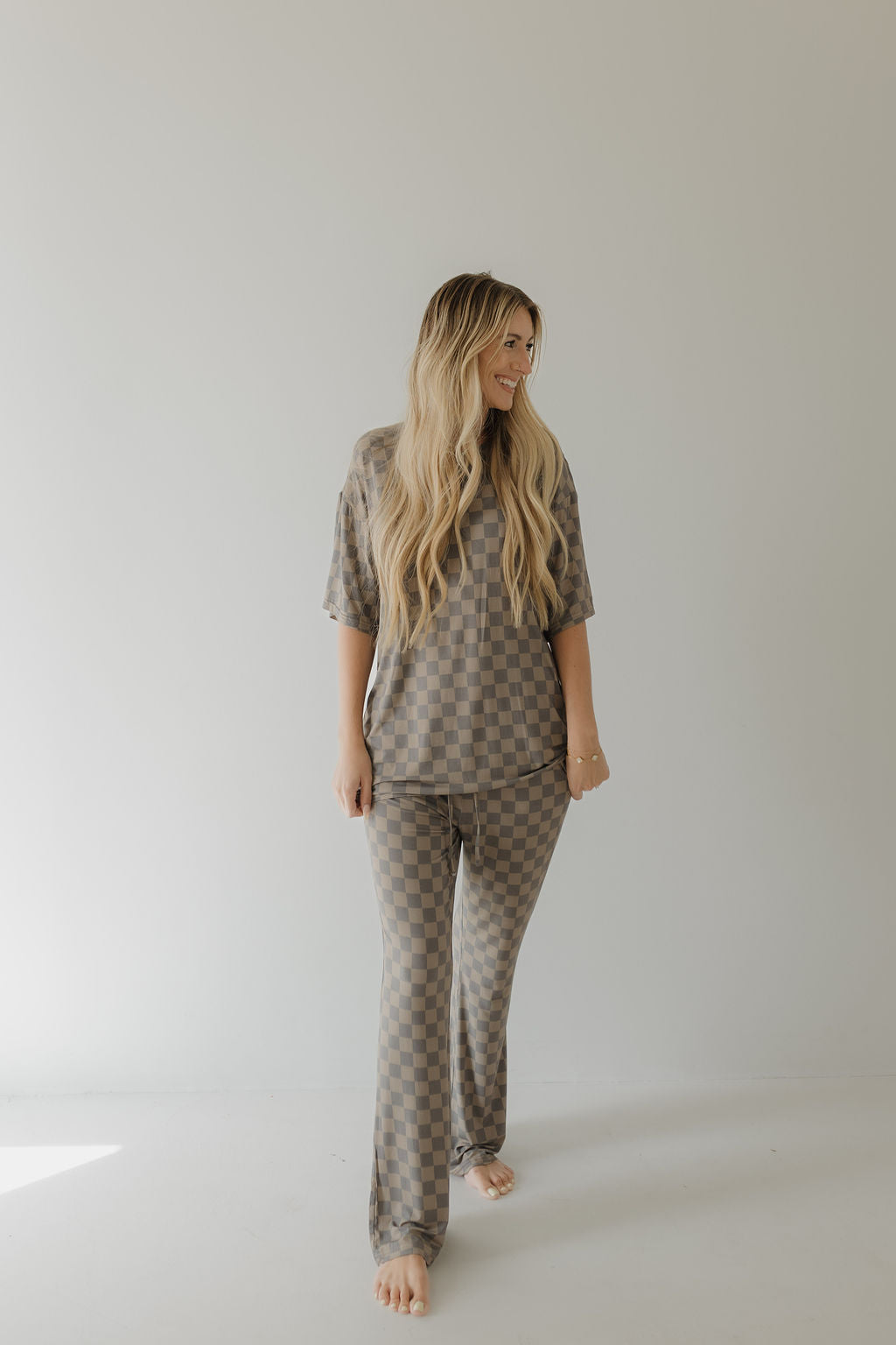 A person with long wavy hair stands against a plain backdrop, wearing the forever french baby Short Sleeve Women's Bamboo Pajama in Faded Checkerboard. The outfit features a breathable bamboo short-sleeved top and long pants. They are smiling to the side, barefoot on the floor.