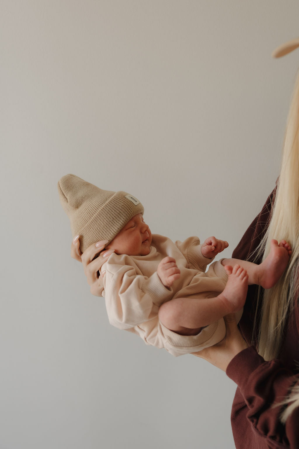 An adult holds a newborn in the forever french baby Sweatshirt Romper | Cashew, paired with a knit hat. The baby's eyes are closed, profile visible, against a plain light background. Only part of the adult and baby can be seen.