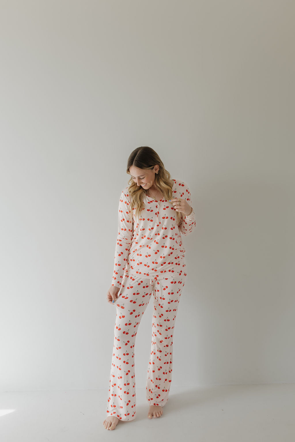A woman wearing the "Women's Bamboo Pajama | Cherry Orchard" by forever french baby stands against a light background, smiling down. Her wavy hair falls over her shoulders as she holds her pants, illustrating year-round comfort.