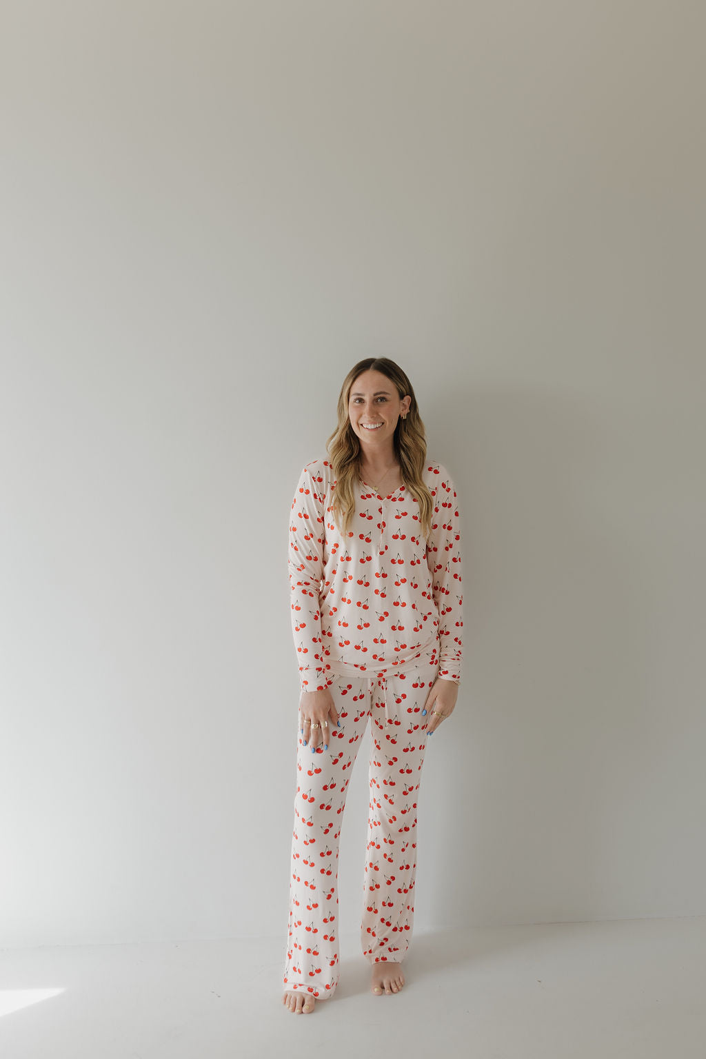 A person with long, light brown hair smiles joyfully, standing barefoot against a plain backdrop in forever french baby's Women's Bamboo Pajama | Cherry Orchard, showcasing the year-round comfy hypo-allergenic fabric with its charming red apple pattern.