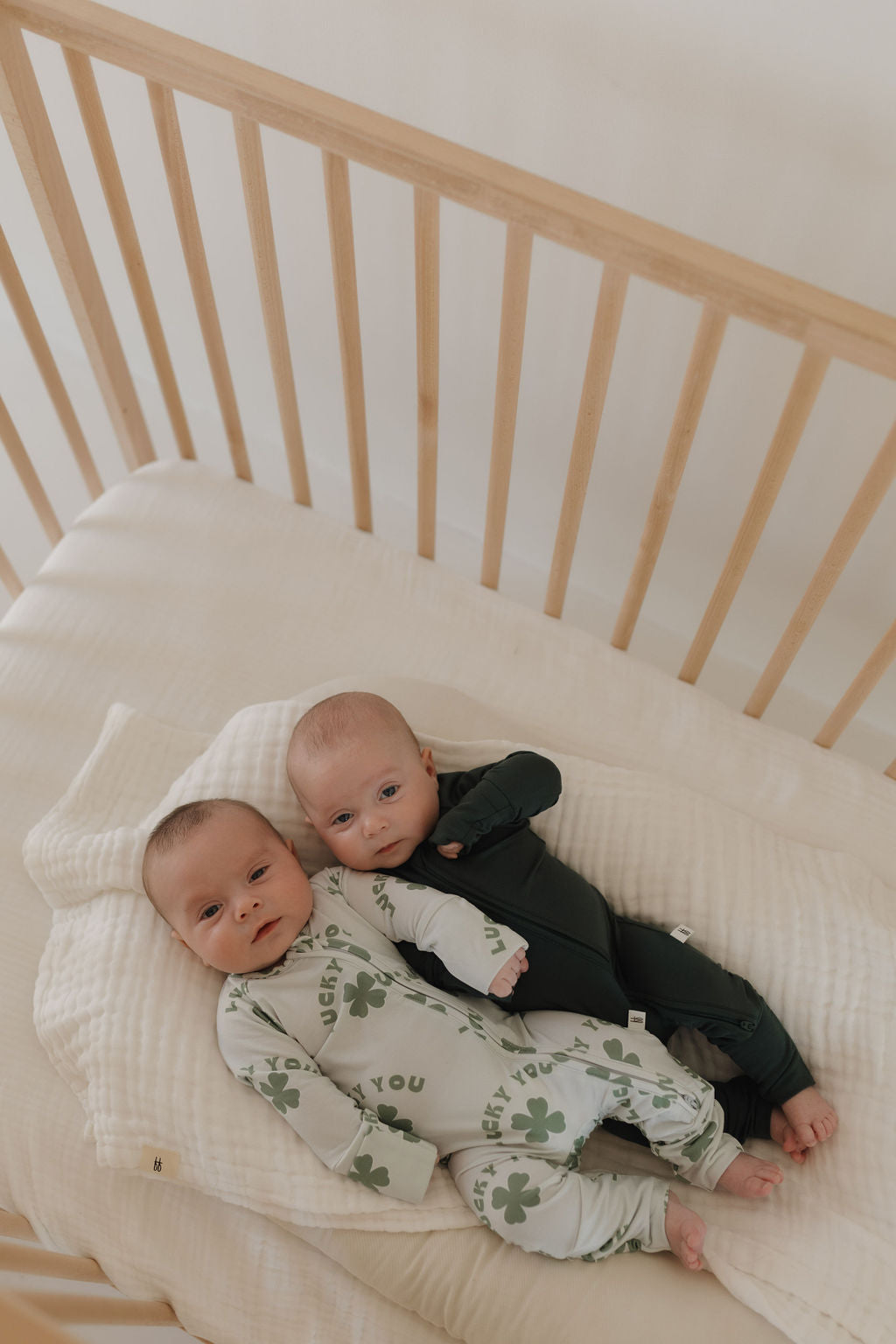 In a wooden crib, two babies rest peacefully wrapped in hypo-allergenic clothing. One wears an emerald green ensemble called Bamboo Zip Pajamas by forever french baby, while the other dons a green outfit with clover patterns on a cream-colored blanket.