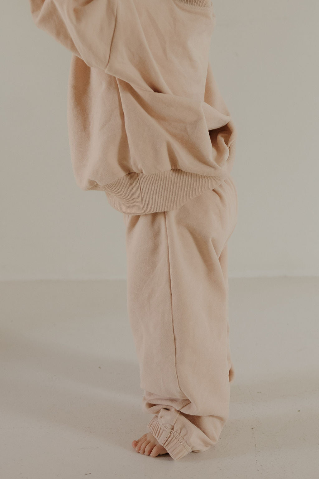 A child wears the forever french baby Child Sweat Set in Cashew—loose, beige sweatpants and a matching sweatshirt—standing barefoot on a light floor. Their face and upper body aren’t visible in this candid moment.
