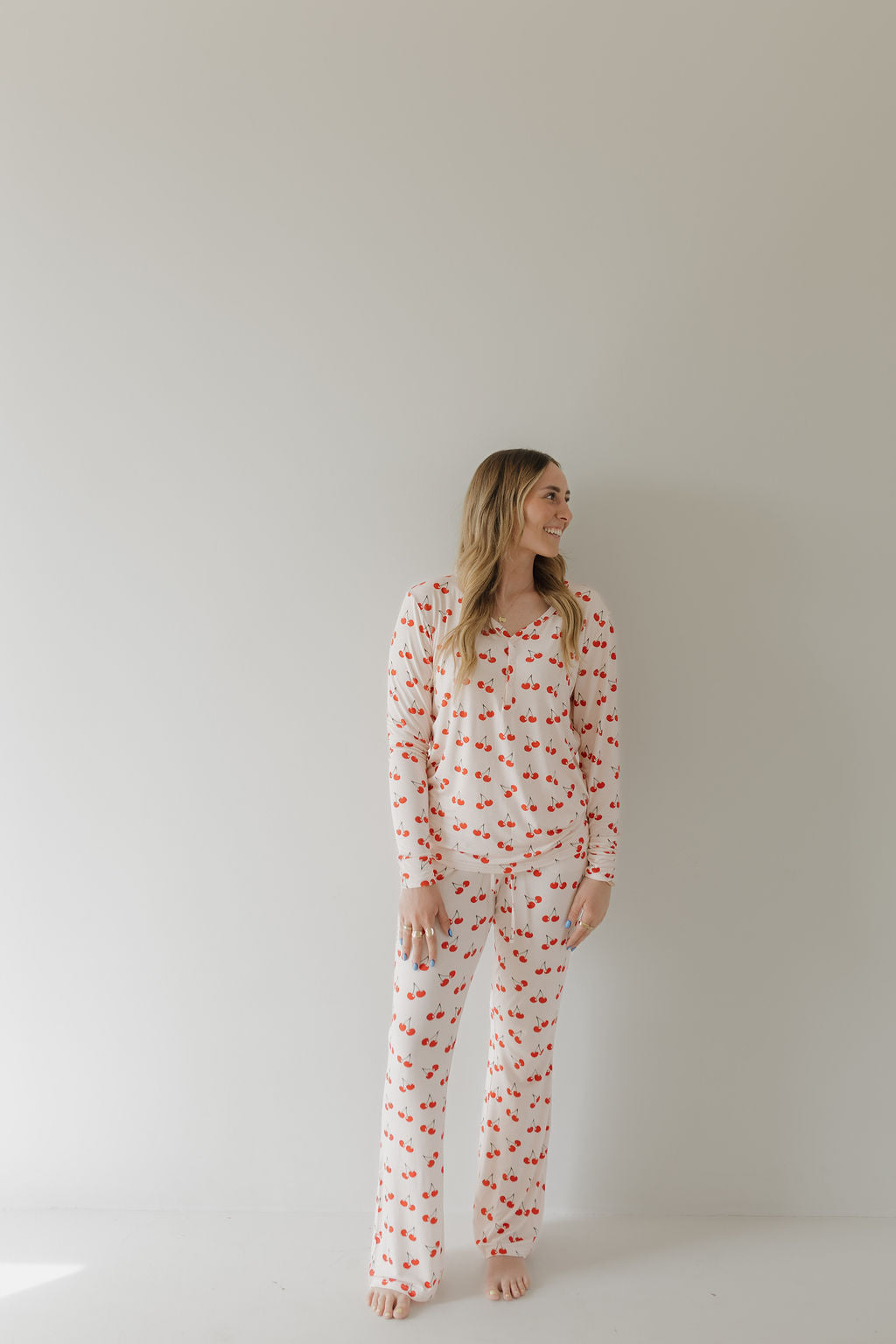 A person with long hair smiles against a plain wall, wearing forever french baby's Women's Bamboo Pajama | Cherry Orchard. Barefoot, they enjoy the hypoallergenic and year-round comfort as their hands rest by their sides.