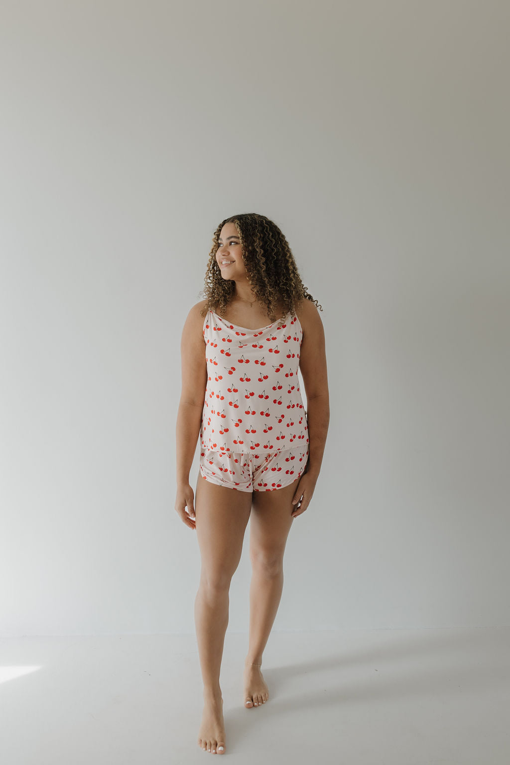 Against a plain backdrop, a person stands barefoot in loose, curly hair wearing the Cami Women's Bamboo Set | Cherry Orchard by forever french baby. This matching sleeveless top and shorts set features a red lips print and is crafted from breathable bamboo fabric under soft diffused lighting.