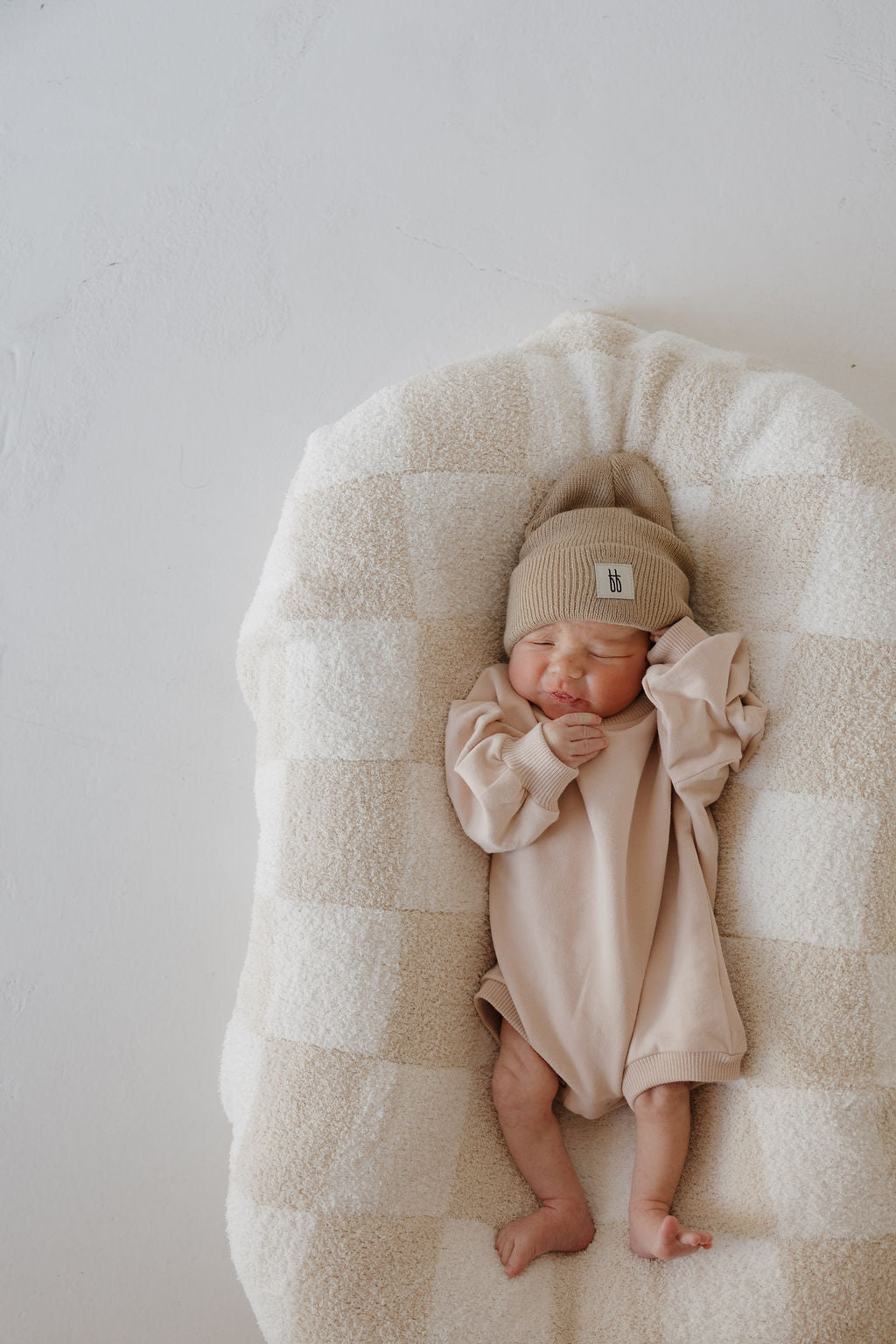 A newborn rests peacefully on a cream and beige checkered blanket, wearing the forever french baby Sweatshirt Romper | Cashew and a beige knit hat, with one hand gently near their face.
