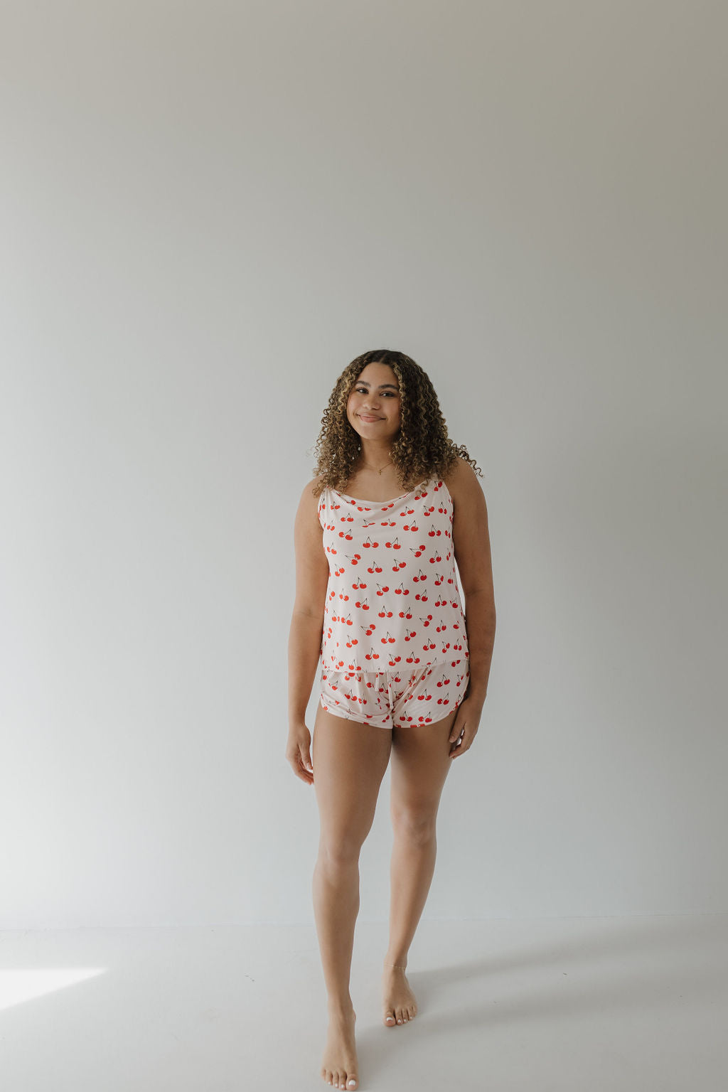 A curly-haired person stands barefoot on a plain, light floor wearing the Cami Women's Bamboo Set by forever french baby. The breathable pajama is hypo-allergenic and features red heart patterns against a minimalist light gray background in the Cherry Orchard design.