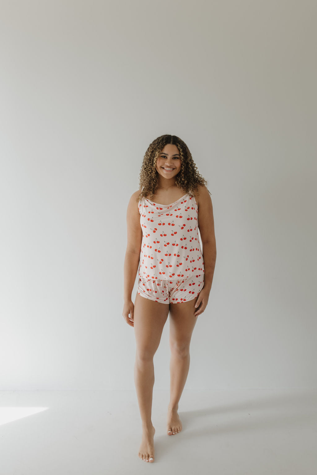 A person with curly hair is smiling, standing barefoot on a light background and enjoying the breathable comfort of their Cami Women's Bamboo Set in Cherry Orchard from forever french baby.