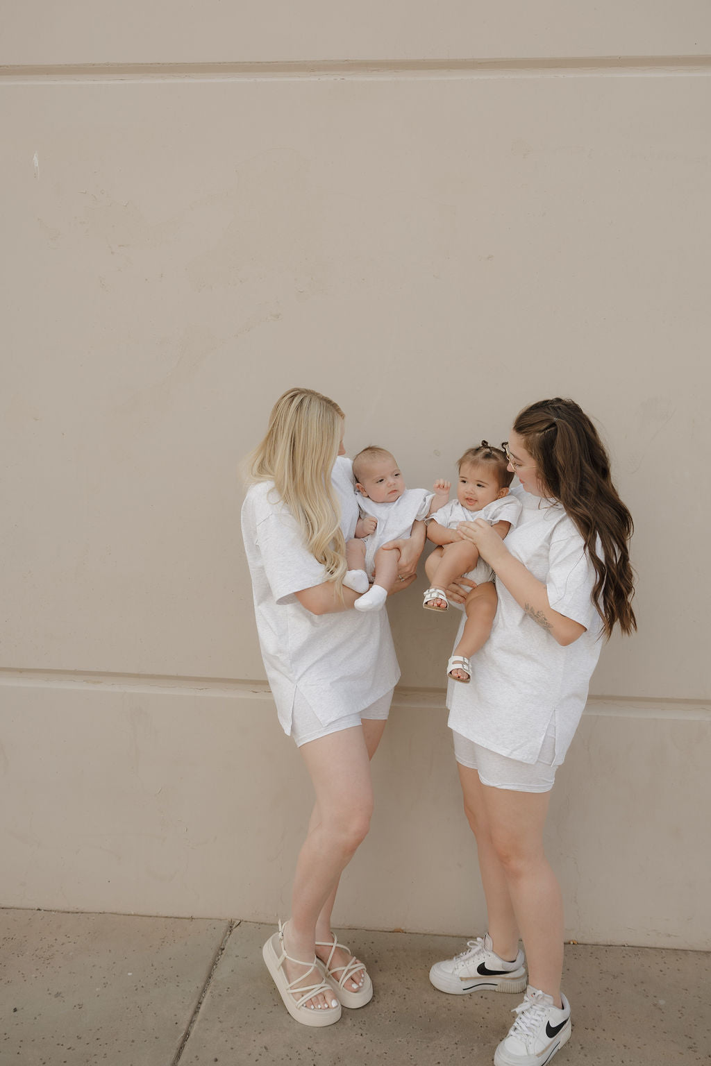 Two women wearing forever french baby Women's Short Set in Heather stand against a beige wall, each holding a baby in white outfits. The women smile at each other while the babies, also facing one another, share the moment.