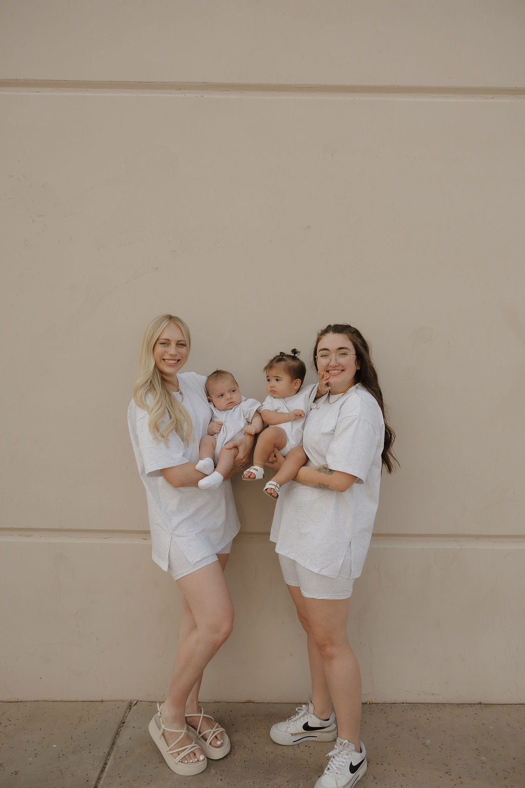 Two women in matching light outfits stand against a beige wall, each holding a baby dressed in the forever french baby Short Sleeve Romper | Heather. Both women and their babies are smiling at the camera.