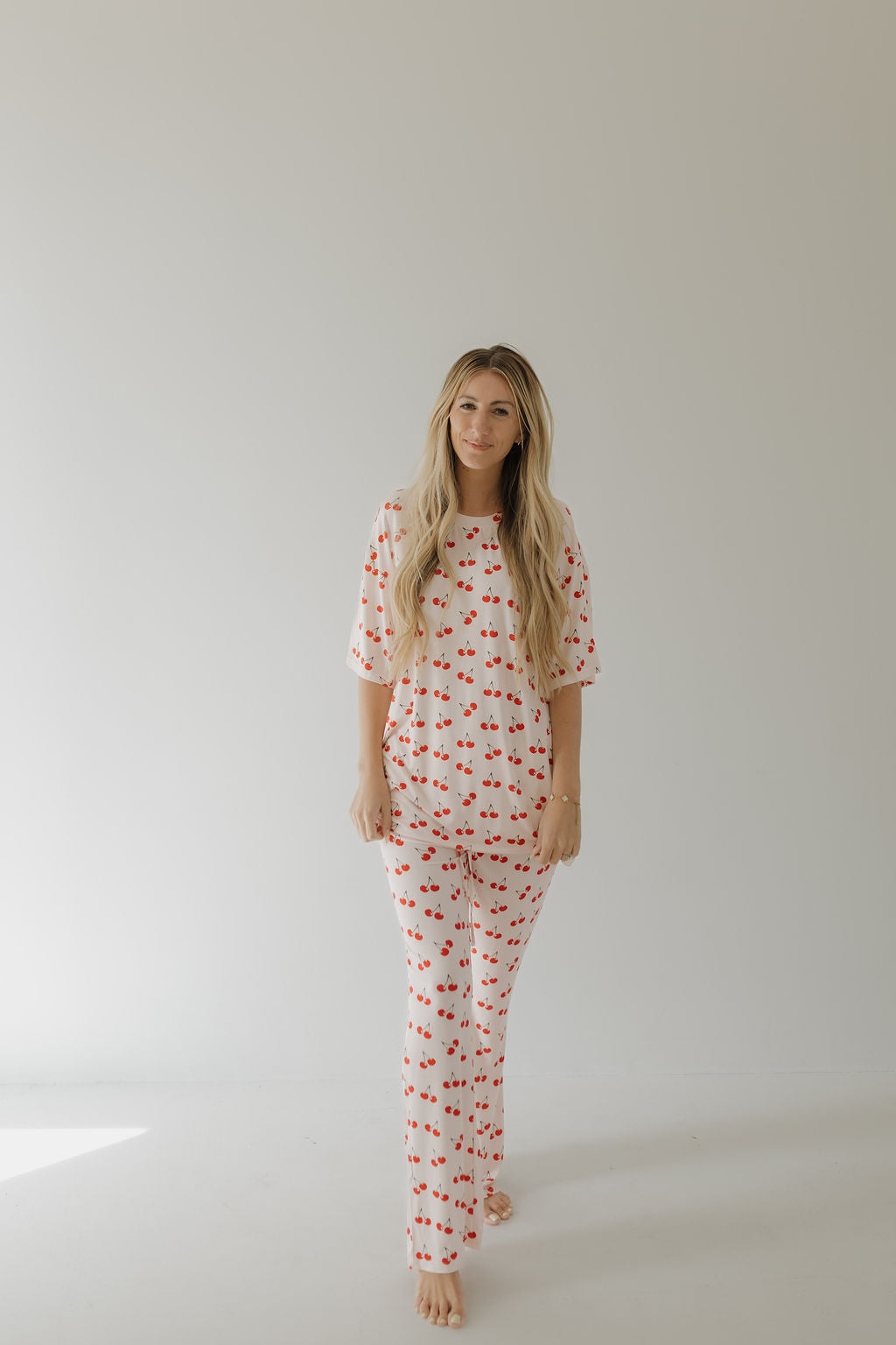 A woman with long blonde hair poses barefoot against a plain background, wearing forever french baby's oversized Short Sleeve Women's Bamboo Pajamas in the Cherry Orchard pattern, which ensures comfort as she looks at the camera.