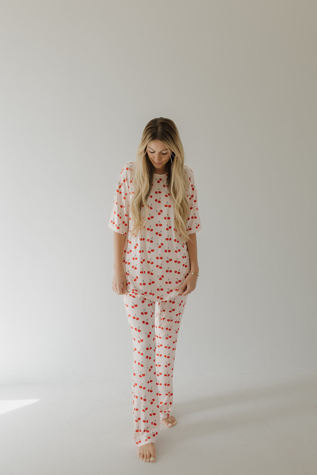 A person with long hair poses against a plain backdrop in an oversized Short Sleeve Women's Bamboo Pajamas by forever french baby, featuring a Cherry Orchard pattern. Their head tilts slightly downward, while hands gently touch the sides of the pajamas.